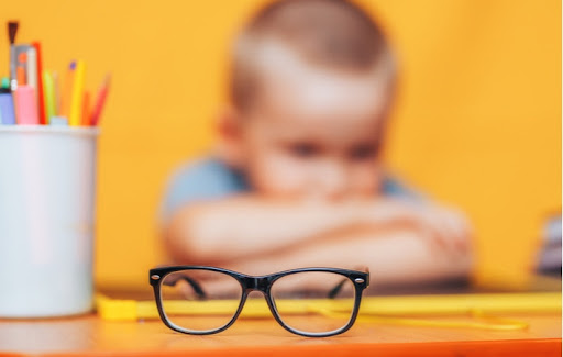 A pair of glasses on a school desk with a blurred boy sitting in the background, highlighting his vision problem's