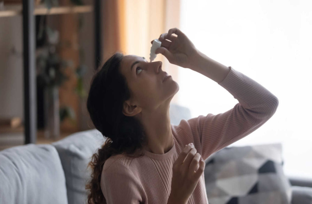 A woman sitting on her couch looking up while applying eye drops to her eye.