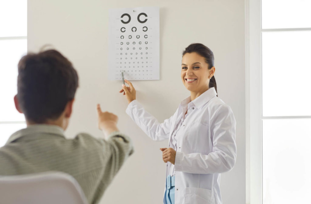 A male child is sitting on a chair from a distance of an optometrist that is standing near a wall while she is pointing at an object in the Snellen chart.