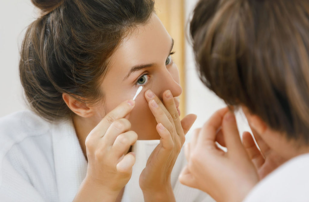 A woman looking in a mirror while she cleans her eyelids with a cotton swab